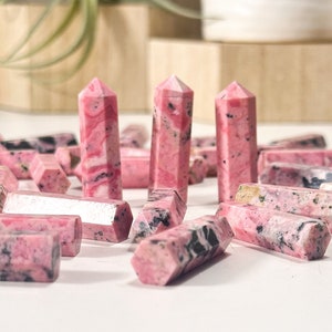May include: A collection of pink and black rhodochrosite crystal points. The crystals are in various shapes and sizes, and some have a natural, rough texture.