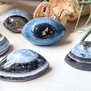May include: A collection of polished, eye-shaped cabochons featuring deep blue and black banded patterns. These smooth, oval-shaped stones display concentric layers of dark colour with lighter blue centres, resembling stylised eyes.