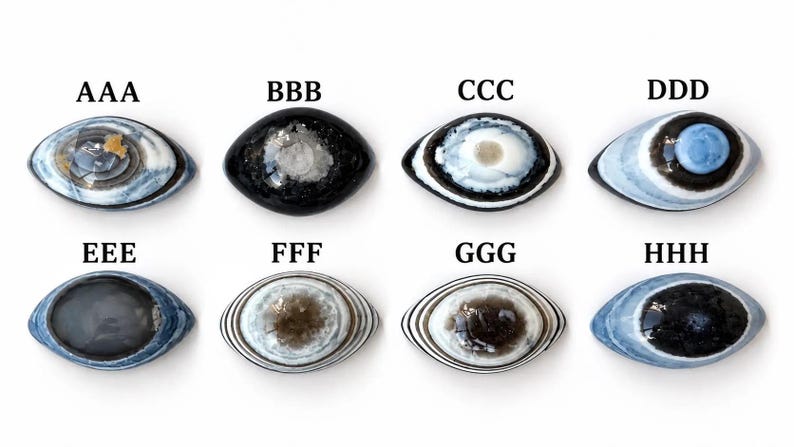 May include: Eight oval-shaped cabochons arranged in two rows, labelled AAA through HHH. Each piece features distinct patterns including concentric rings, dark centres, and blue, white, and black colour variations. The stones have a smooth, polished surface with varied textures and colour gradients throughout the collection.