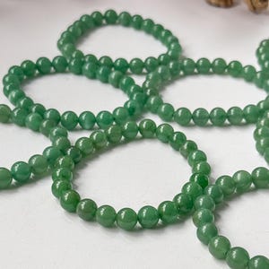 Puede incluir: Primer plano de varios hilos de cuentas de piedras preciosas verdes. Las cuentas redondas y pulidas son de un color verde vibrante. Est&aacute;n dispuestas sobre una superficie blanca. La imagen est&aacute; bien iluminada.