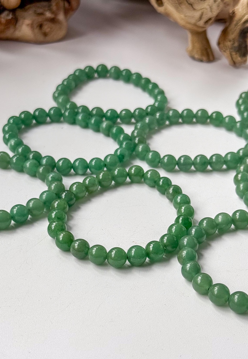 Puede incluir: Primer plano de varios hilos de cuentas de piedras preciosas verdes. Las cuentas redondas y pulidas son de un color verde vibrante. Est&aacute;n dispuestas sobre una superficie blanca. La imagen est&aacute; bien iluminada.