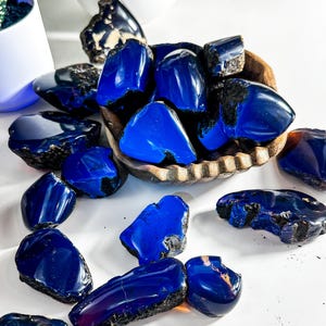 Puede incluir: Una colecci&oacute;n de piedras de &aacute;mbar azul, algunas en un cuenco de madera, otras esparcidas sobre una superficie blanca. Las piedras son de varios tama&ntilde;os y formas, con un color azul intenso y una apariencia ligeramente nublada.
