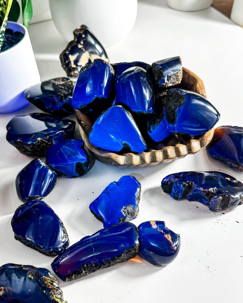 Puede incluir: Una colecci&oacute;n de piedras de &aacute;mbar azul, algunas en un cuenco de madera, otras esparcidas sobre una superficie blanca. Las piedras son de varios tama&ntilde;os y formas, con un color azul intenso y una apariencia ligeramente nublada.