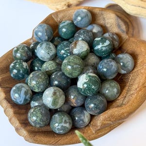 May include: A collection of moss agate spheres in a wooden bowl. The spheres are a mix of green, white, and gray, with a marbled appearance. The bowl is made of light brown wood. The background includes a piece of driftwood and a green plant.