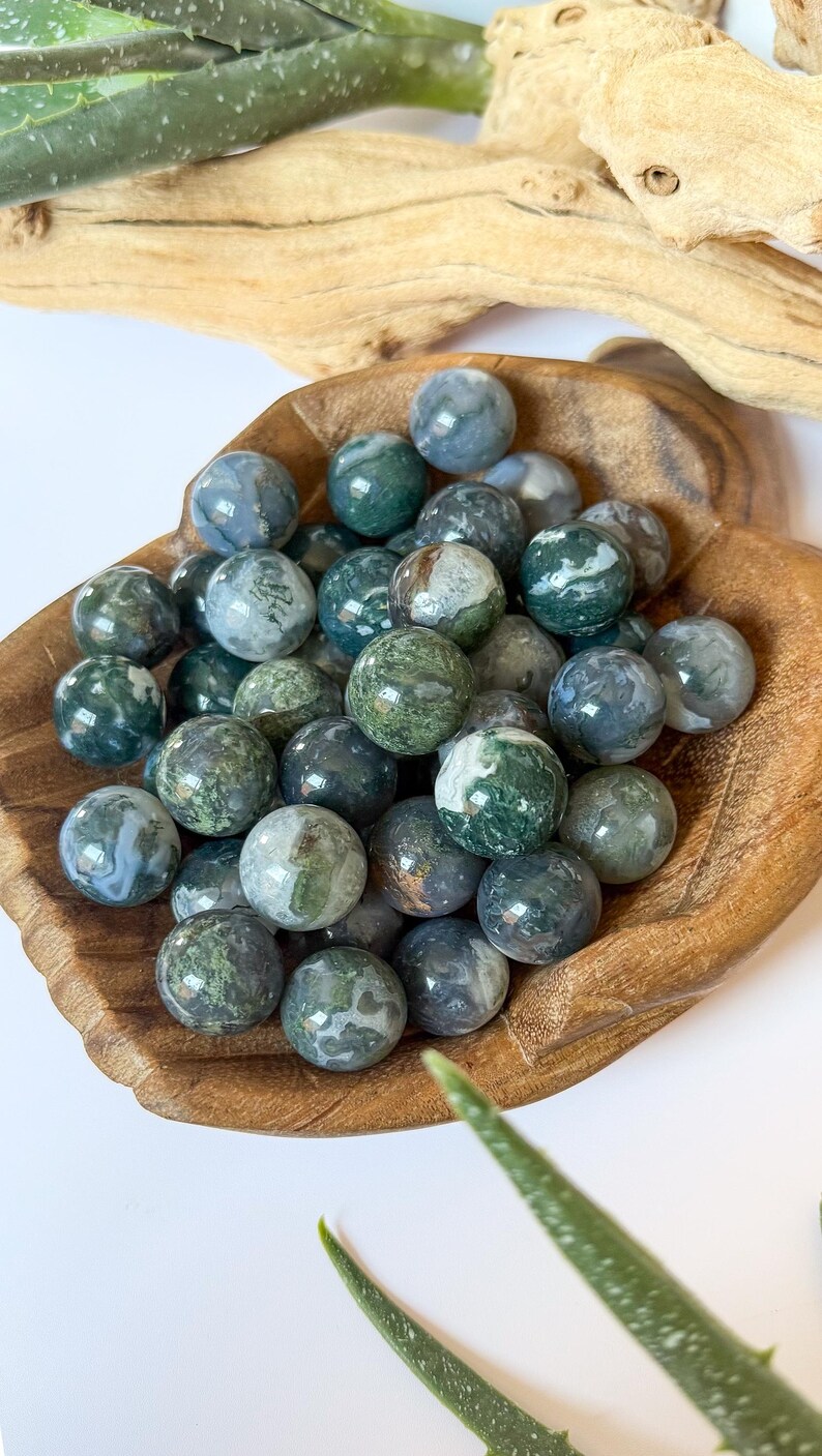 May include: A collection of moss agate spheres in a wooden bowl. The spheres are a mix of green, white, and gray, with a marbled appearance. The bowl is made of light brown wood. The background includes a piece of driftwood and a green plant.