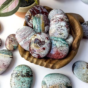 May include: A collection of polished, oval-shaped ocean jasper stones in a wooden bowl. The stones feature a variety of colours including deep red, forest green, cream, and grey, with intricate swirling patterns, banding, and small dark speckles.