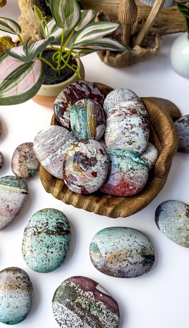 May include: A collection of polished, oval-shaped ocean jasper stones in a wooden bowl. The stones feature a variety of colours including deep red, forest green, cream, and grey, with intricate swirling patterns, banding, and small dark speckles.