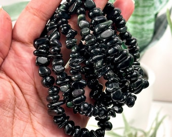 May include: A strand of black obsidian chip beads, with a slightly iridescent sheen, is shown in a hand. The beads are small and irregular in shape, and they are strung together on a clear elastic cord.