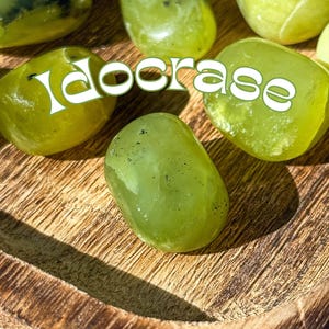 Op de afbeelding: Een verzameling gladde, groene Idocrase-tumbled stenen op een houten oppervlak. De stenen zijn van verschillende groottes en vormen, met een lichtgroene kleur en enkele donkergroene markeringen.