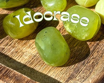May include: A collection of smooth, green Idocrase tumbled stones on a wooden surface. The stones are various sizes and shapes, with a light green color and some darker green markings.