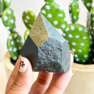 May include: A dark gray, faceted crystal point with a rough, textured surface. The crystal is being held in a hand with brown floral nail art.