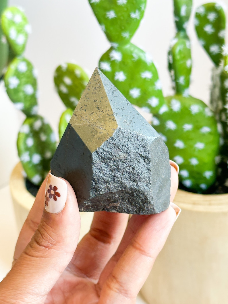 May include: A dark gray, faceted crystal point with a rough, textured surface. The crystal is being held in a hand with brown floral nail art.