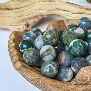 May include: A wooden bowl filled with polished, round gemstones. The stones are primarily green and blue with mottled patterns. The bowl is made of light brown wood and sits on a white surface. A piece of driftwood is in the background.