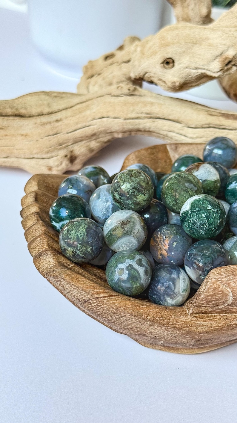 May include: A wooden bowl filled with polished, round gemstones. The stones are primarily green and blue with mottled patterns. The bowl is made of light brown wood and sits on a white surface. A piece of driftwood is in the background.