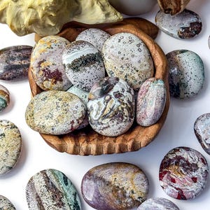 May include: A collection of polished, oval-shaped ocean jasper stones in a carved wooden bowl, surrounded by loose stones on a white surface. The stones feature varied patterns of green, brown, cream, and red with speckled and banded textures.