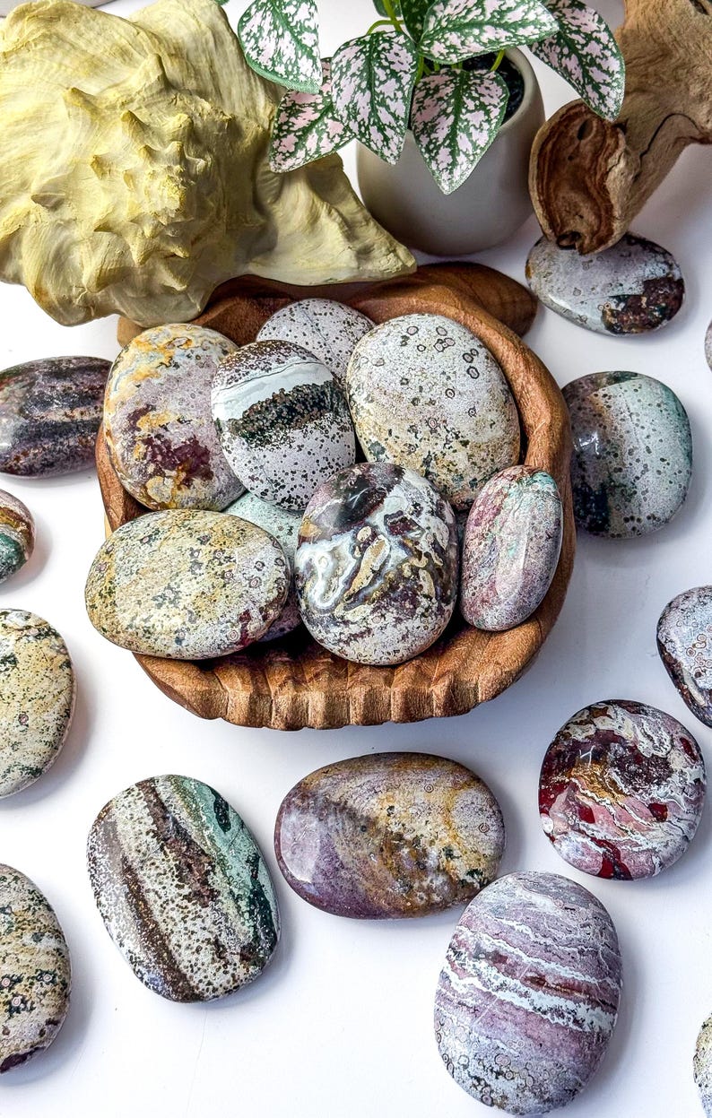 May include: A collection of polished, oval-shaped ocean jasper stones in a carved wooden bowl, surrounded by loose stones on a white surface. The stones feature varied patterns of green, brown, cream, and red with speckled and banded textures.