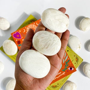 May include: Three white, smooth, oval-shaped stones with a slightly textured surface. The stones are arranged in a hand, with the largest stone in the center.