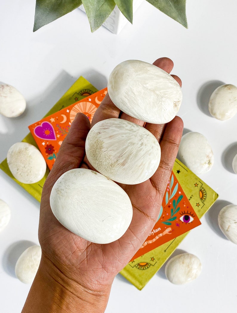 May include: Three white, smooth, oval-shaped stones with a slightly textured surface. The stones are arranged in a hand, with the largest stone in the center.
