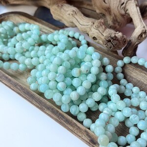 May include: Strands of light blue gemstone beads are displayed in a wooden tray. The beads are round and have a slightly marbled appearance. The tray is rectangular and has a natural wood grain. A piece of driftwood is in the background.