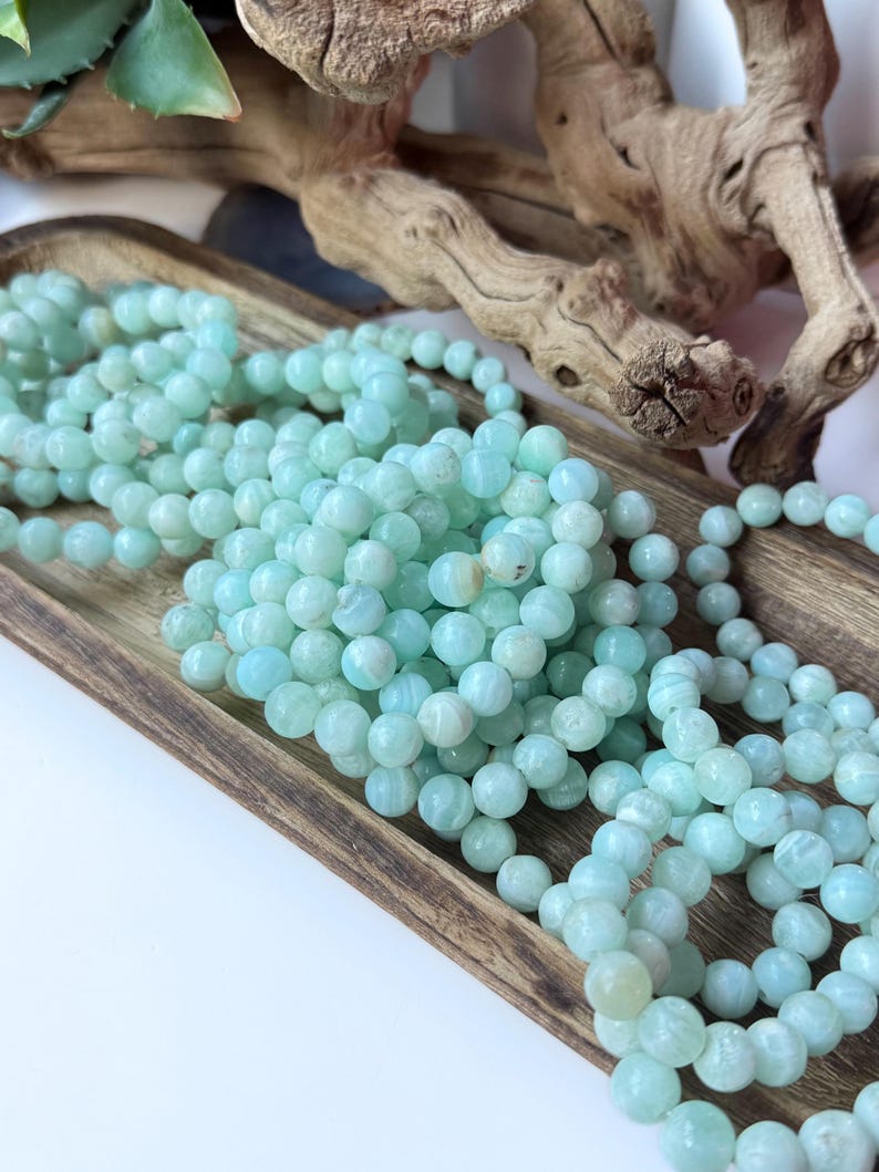 May include: Strands of light blue gemstone beads are displayed in a wooden tray. The beads are round and have a slightly marbled appearance. The tray is rectangular and has a natural wood grain. A piece of driftwood is in the background.