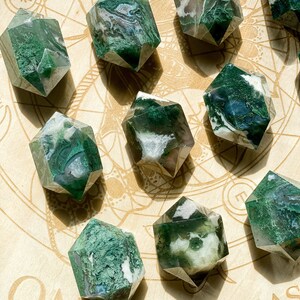 May include: A selection of faceted moss agate crystal points. The crystals display clear, white, and green hues, with detailed patterns. They are arranged on a light-coloured wooden surface with etched designs.