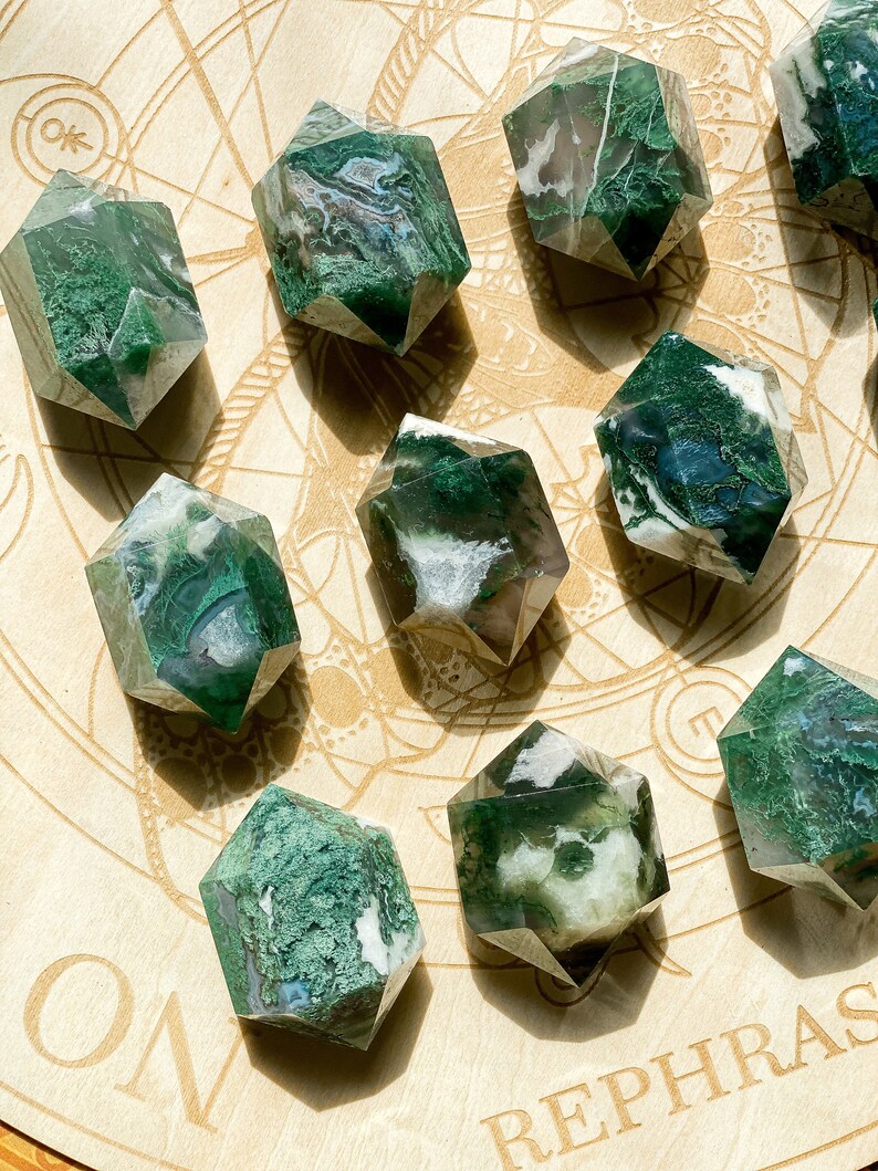 May include: A selection of faceted moss agate crystal points. The crystals display clear, white, and green hues, with detailed patterns. They are arranged on a light-coloured wooden surface with etched designs.