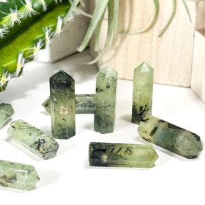 May include: A collection of seven green and black crystal points. The points are all different sizes and shapes, and they are arranged in a random pattern on a white surface.