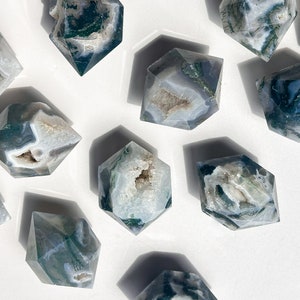 May include: A selection of hexagonal crystal stones. The stones are white and grey with green, mossy inclusions. Some have a hollowed-out centre. The crystals are displayed on a white surface.