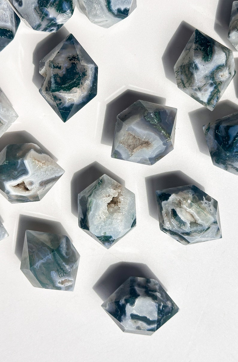 May include: A selection of hexagonal crystal stones. The stones are white and grey with green, mossy inclusions. Some have a hollowed-out centre. The crystals are displayed on a white surface.