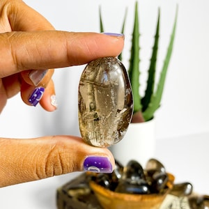 May include: A smooth, oval-shaped, light brown smoky quartz crystal. The crystal is being held by a hand with purple nail polish. The crystal is in focus, while the background is blurred.