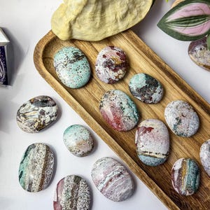May include: A collection of smooth, polished oval stones featuring a variety of natural patterns and colours including shades of mint green, deep red, white, and brown, arranged on a long, narrow wooden tray with a visible wood grain texture.