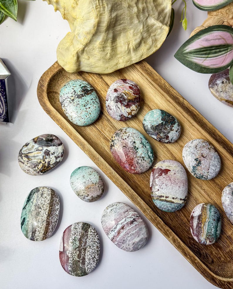 May include: A collection of smooth, polished oval stones featuring a variety of natural patterns and colours including shades of mint green, deep red, white, and brown, arranged on a long, narrow wooden tray with a visible wood grain texture.