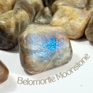 以下が含まれることがあります： 研磨されたベロモライトムーンストーンのグループのクローズアップ。石は薄い茶色で、青い虹色の輝きがあります。石は滑らかで磨かれています。