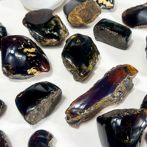 Puede incluir: Una colecci&oacute;n de 14 piedras de &aacute;mbar negro en bruto y natural. Las piedras son de varios tama&ntilde;os y formas, algunas tienen un ligero tono p&uacute;rpura.