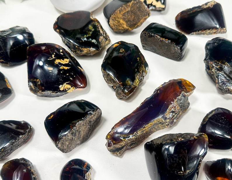 Puede incluir: Una colecci&oacute;n de 14 piedras de &aacute;mbar negro en bruto y natural. Las piedras son de varios tama&ntilde;os y formas, algunas tienen un ligero tono p&uacute;rpura.