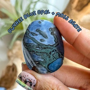 May include: Oval-shaped Owyhee Blue Opal and Moss Agate stone. The stone features a swirling pattern of blue and green hues. The text "OWYHEE BLUE OPAL + MOSS AGATE" is written in a light blue font.