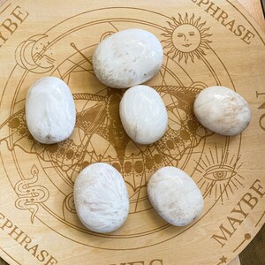 May include: Six white, smooth, oval-shaped stones on a wooden board with a circular design and the words "YES", "REPHRASE", "MAYBE", and "YES" printed on it.
