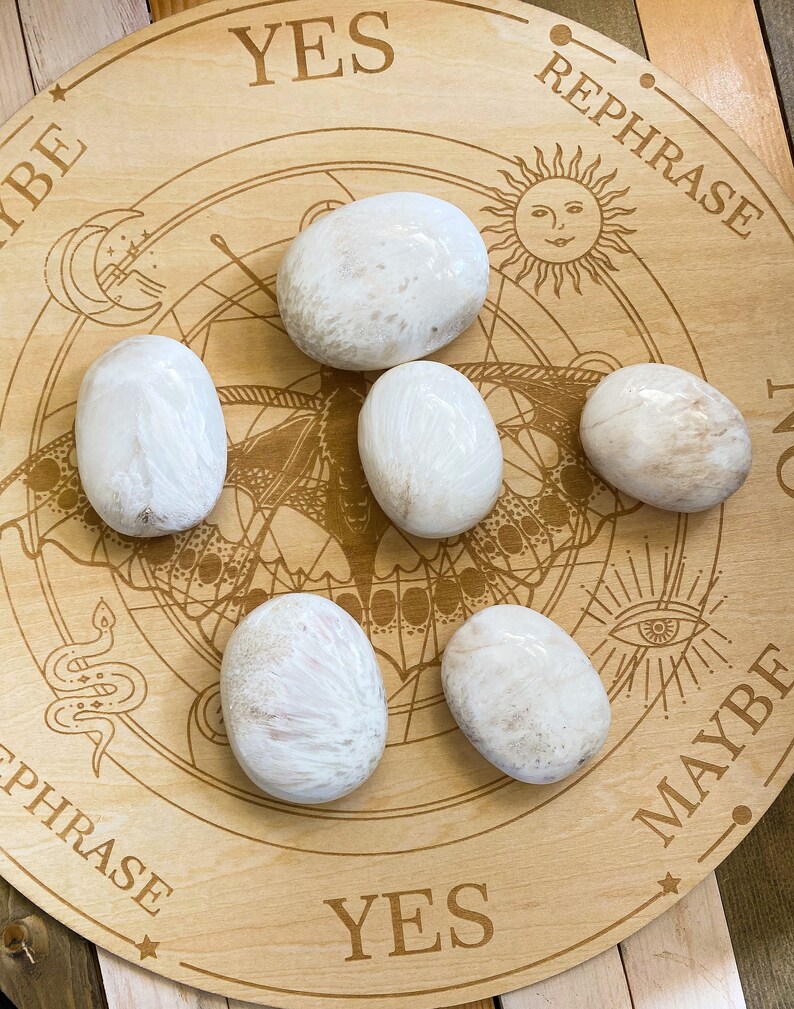May include: Six white, smooth, oval-shaped stones on a wooden board with a circular design and the words "YES", "REPHRASE", "MAYBE", and "YES" printed on it.