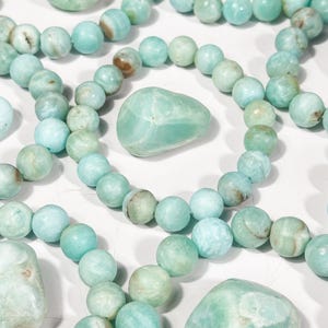 May include: An overhead shot showcases multiple strands of light blue-green beaded bracelets and several polished stones. The beads are round, and the stones are smooth and irregular in shape. The color palette is soft and natural, with variations in tone.