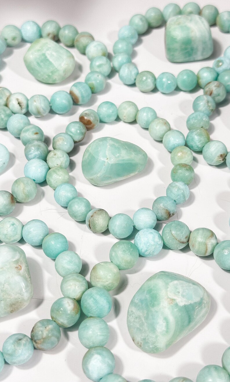May include: An overhead shot showcases multiple strands of light blue-green beaded bracelets and several polished stones. The beads are round, and the stones are smooth and irregular in shape. The color palette is soft and natural, with variations in tone.