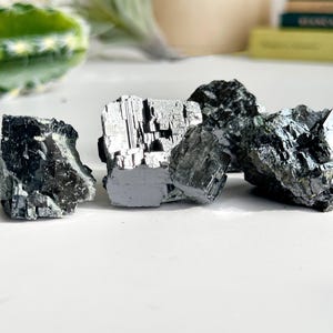 May include: Four raw, unpolished, dark grey and silver pyrite crystals on a white surface.