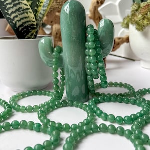 Puede incluir: Un objeto decorativo verde en forma de cactus sostiene varias hebras de collares de cuentas verdes. Las cuentas son peque&ntilde;as y redondas. El cactus es liso y brillante. El fondo incluye plantas en macetas y una superficie blanca.