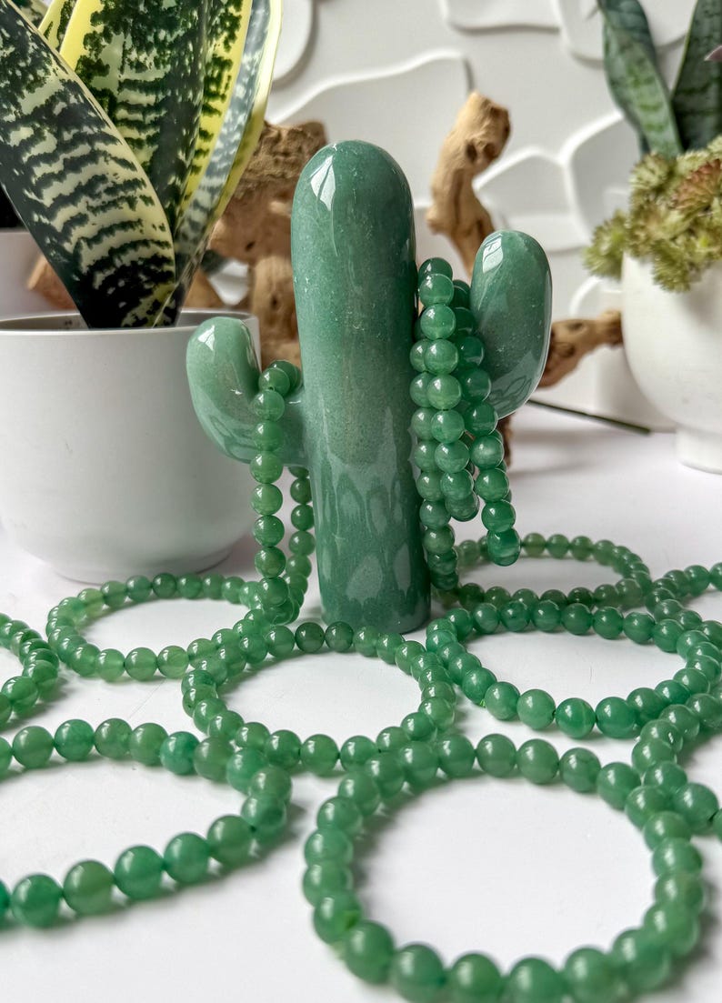 Puede incluir: Un objeto decorativo verde en forma de cactus sostiene varias hebras de collares de cuentas verdes. Las cuentas son peque&ntilde;as y redondas. El cactus es liso y brillante. El fondo incluye plantas en macetas y una superficie blanca.