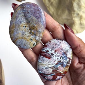 May include: Two polished, oval-shaped stones with smooth surfaces. The top stone features a blend of muted purple, tan, and cream tones with small dark speckles. The bottom stone displays a vibrant mix of deep red, white, and blue-grey patterns.