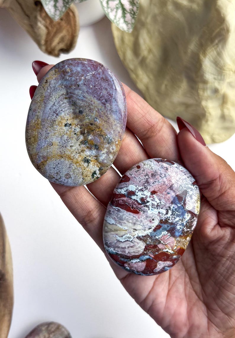 May include: Two polished, oval-shaped stones with smooth surfaces. The top stone features a blend of muted purple, tan, and cream tones with small dark speckles. The bottom stone displays a vibrant mix of deep red, white, and blue-grey patterns.