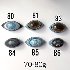 May include: Six polished, eye-shaped stones arranged in two rows, labelled 80, 81, 83, 84, 85, and 86. The stones feature blue, grey, and brown banded patterns. The text 70-80g is printed at the bottom, indicating the weight range of the stones.