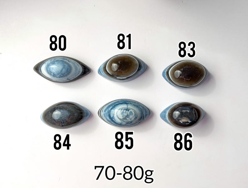 May include: Six polished, eye-shaped stones arranged in two rows, labelled 80, 81, 83, 84, 85, and 86. The stones feature blue, grey, and brown banded patterns. The text 70-80g is printed at the bottom, indicating the weight range of the stones.