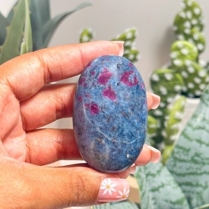 May include: An oval-shaped polished gemstone featuring a deep blue base colour with scattered, vibrant magenta-pink inclusions throughout the surface. The stone has a smooth, glossy texture and exhibits natural mineral variations across its face.