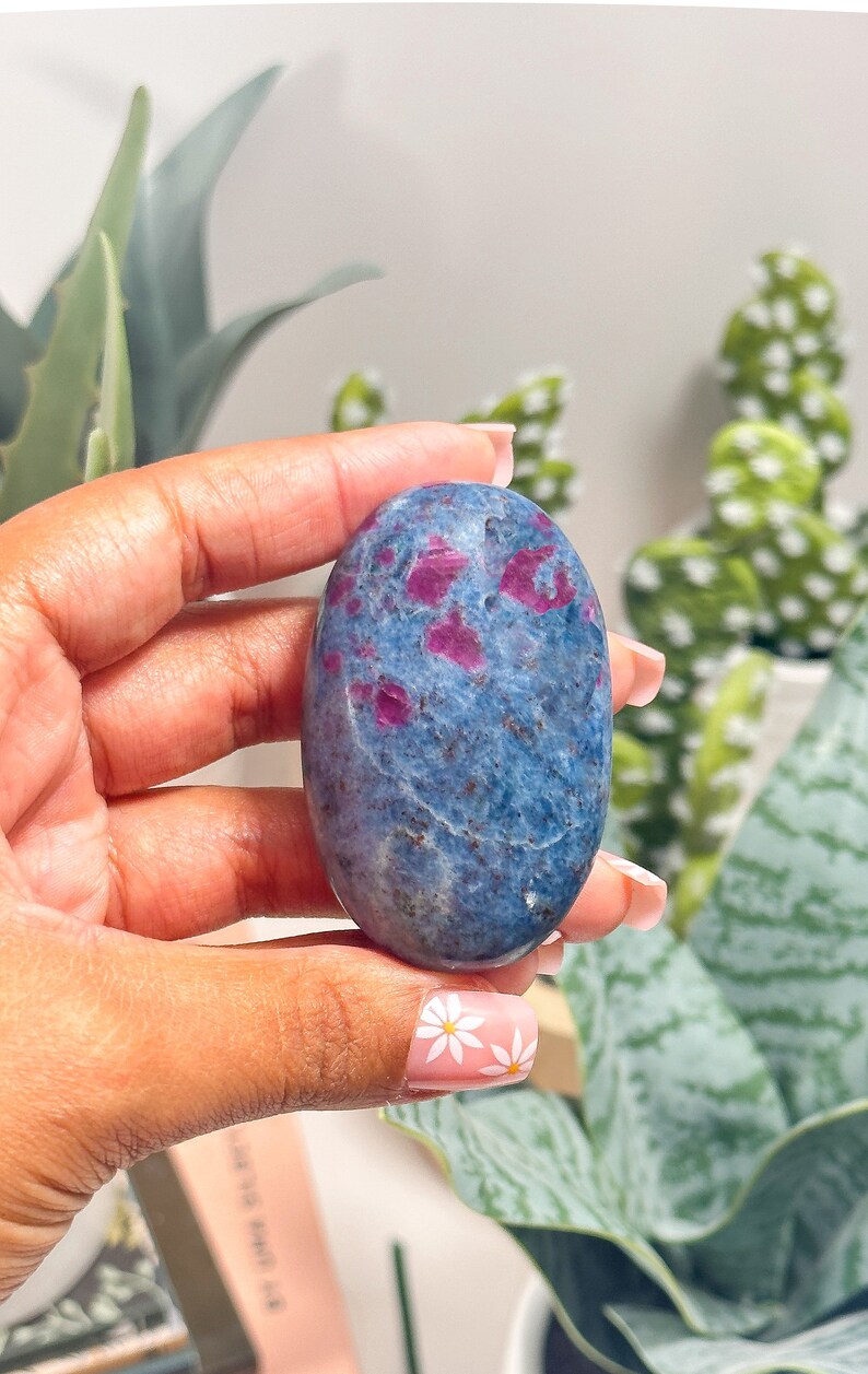 May include: An oval-shaped polished gemstone featuring a deep blue base colour with scattered, vibrant magenta-pink inclusions throughout the surface. The stone has a smooth, glossy texture and exhibits natural mineral variations across its face.
