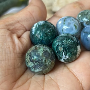 May include: A collection of polished moss agate spheres in varying shades of green and white. The spheres are held in a hand, showcasing their natural patterns and textures. The gemstones have a smooth, rounded shape.